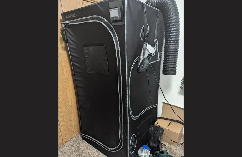 An exterior view of an AC Infinity grow tent with black fabric walls sucking inward due to negative pressure. A digital environmental controller is mounted on the top and black ducting exits the side.