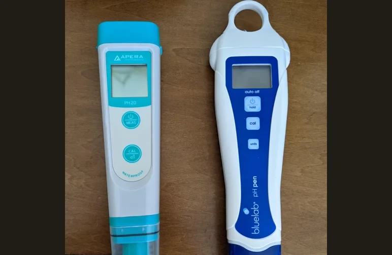 Two professional-grade digital pH pens, an Apera PH20 and a Bluelab pH Pen, lying side-by-side on a wooden surface for cannabis nutrient testing.