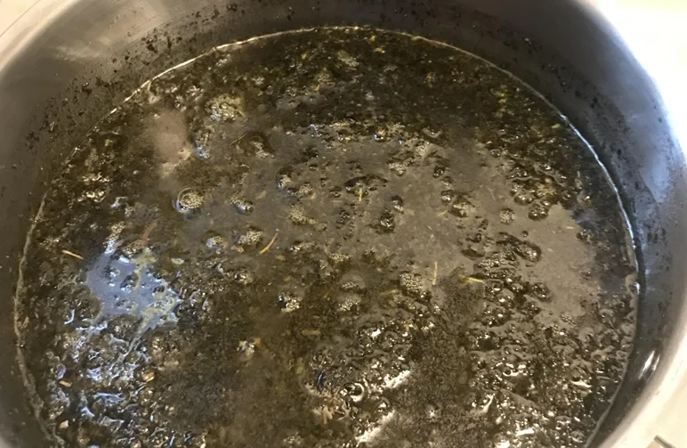 A top-down view of a dark green cannabis, butter, and water mixture simmering gently in a stainless steel saucepan with small bubbles on the surface.