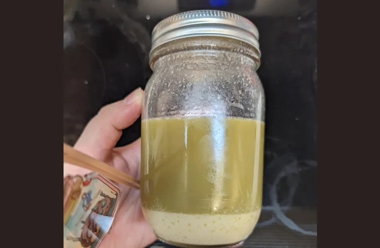 A glass mason jar held by a hand, showing a thick green layer of solid cannabutter on top of a lighter, cloudy layer of water.