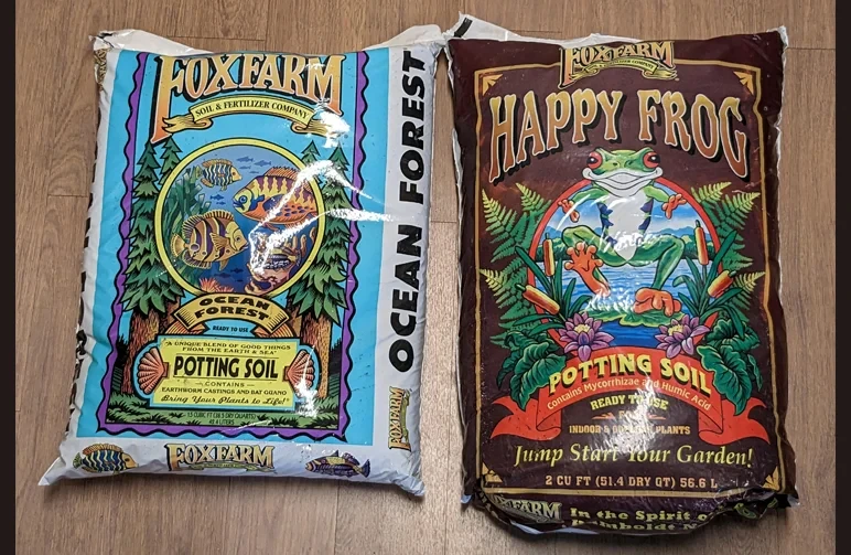 Seitenvergleich von Fox Farm Ocean Forest (blauer Sack) und Happy Frog (brauner Sack) Blumenerden auf einem Holzboden.