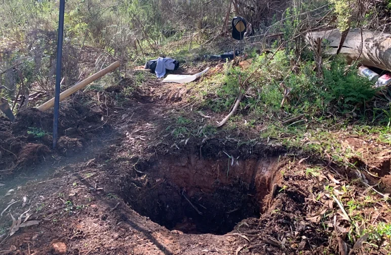Ein tiefes Pflanzloch, gegraben in einheimischen rötlich-braunen Boden in einer Waldlichtung, mit Spitzhacke und Wanderausrüstung im Hintergrund, das die Vorbereitung für einen Guerilla-Cannabis-Grow zeigt.